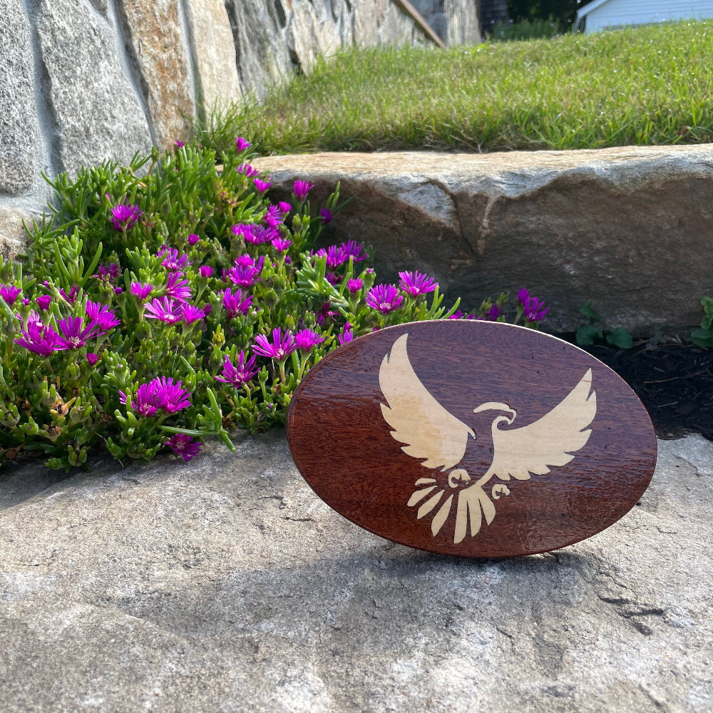 eagle faceing forward with wings outspread inlaid into a wooden hitch cover, balanced on a rock with green grass and purple flowers behind it.