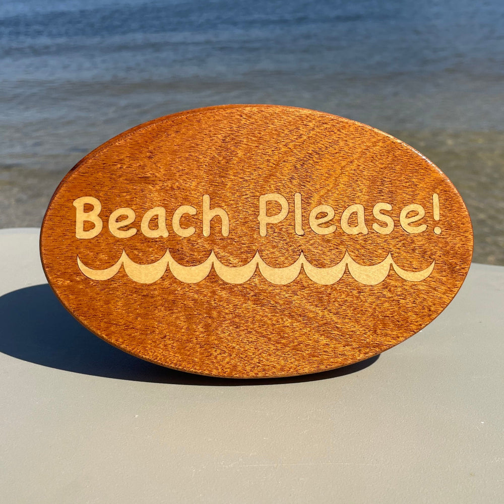 A wooden hitch cover with the phrase 'Beach Please!' engraved on it, placed on a vehicle near a beach.