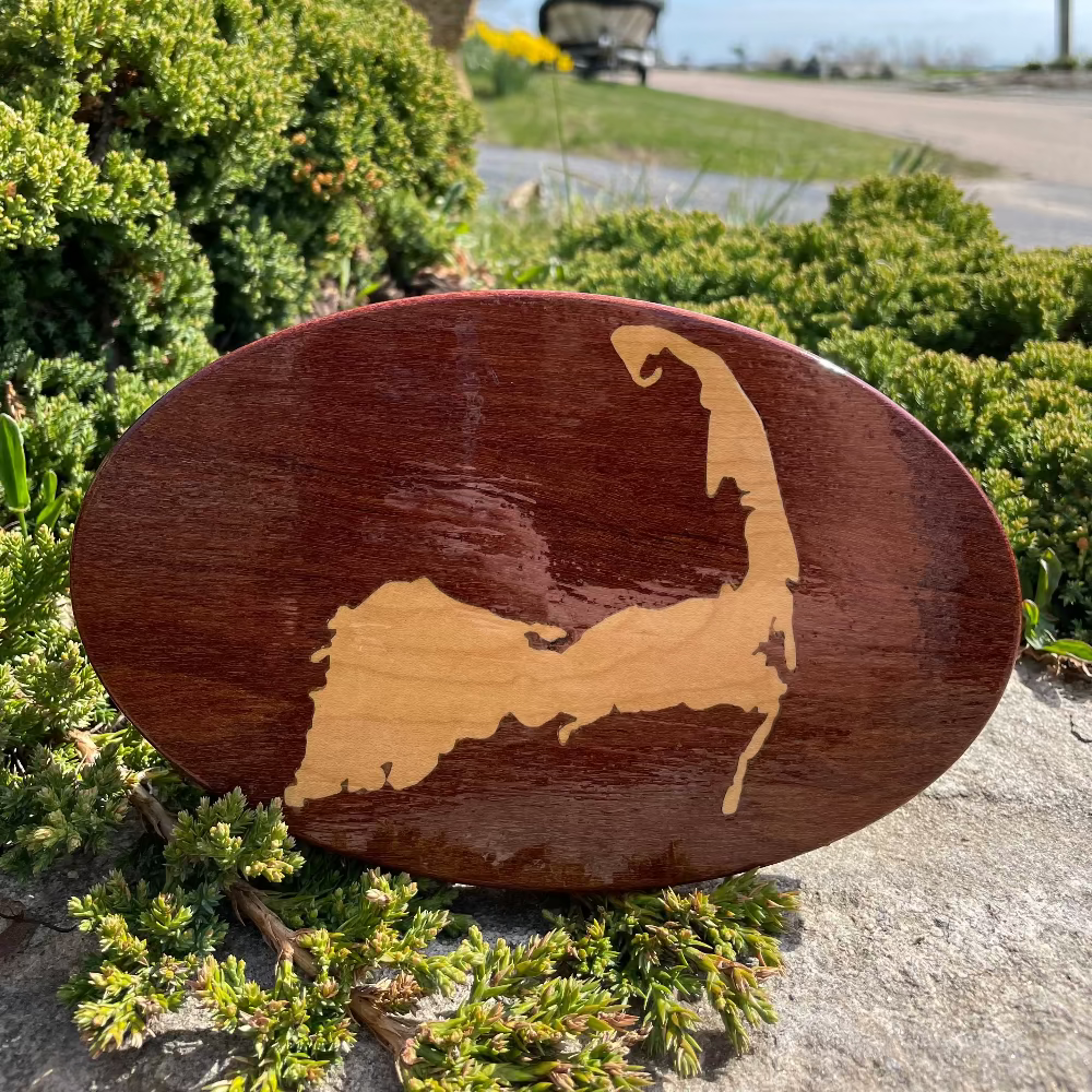 A wooden hitch cover featuring a Cape Cod map design, placed on a stone surface with green plants in the background.