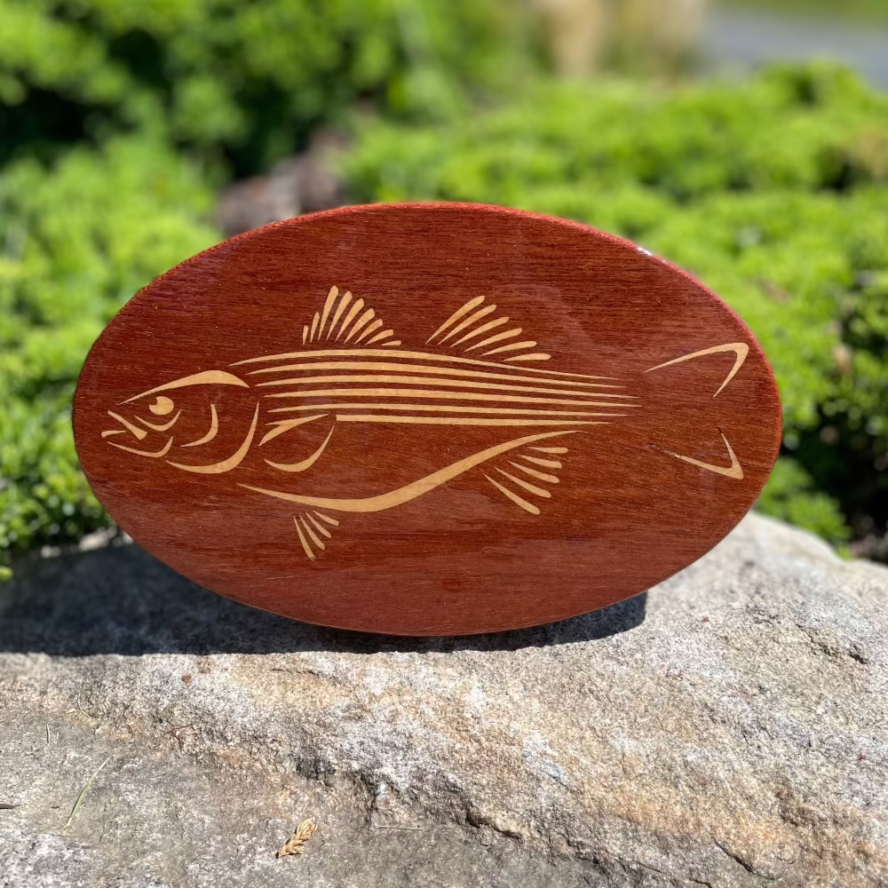 An oval-shaped, artisan crafted wooden hitch-cover with a Cherokee Striped Bass design, placed on a rock.