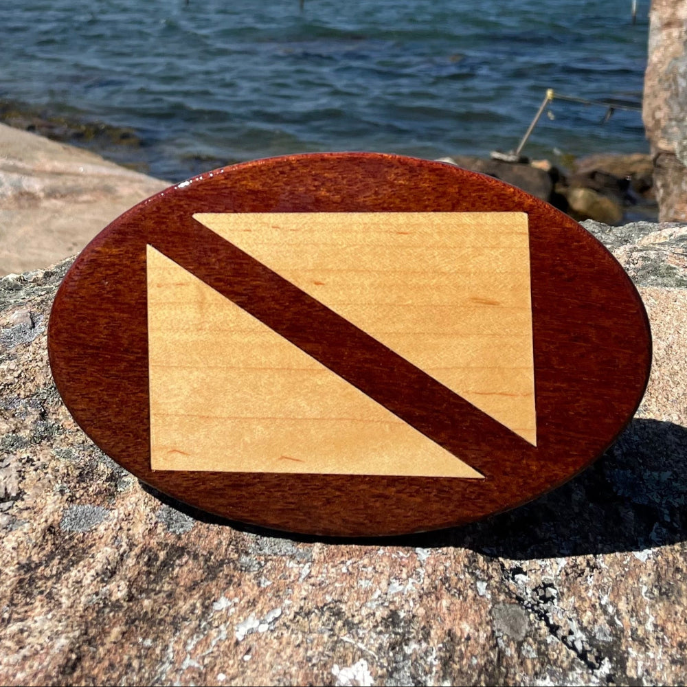 A wooden hitch cover with a flag design, placed on a rock near a body of water and a pier.