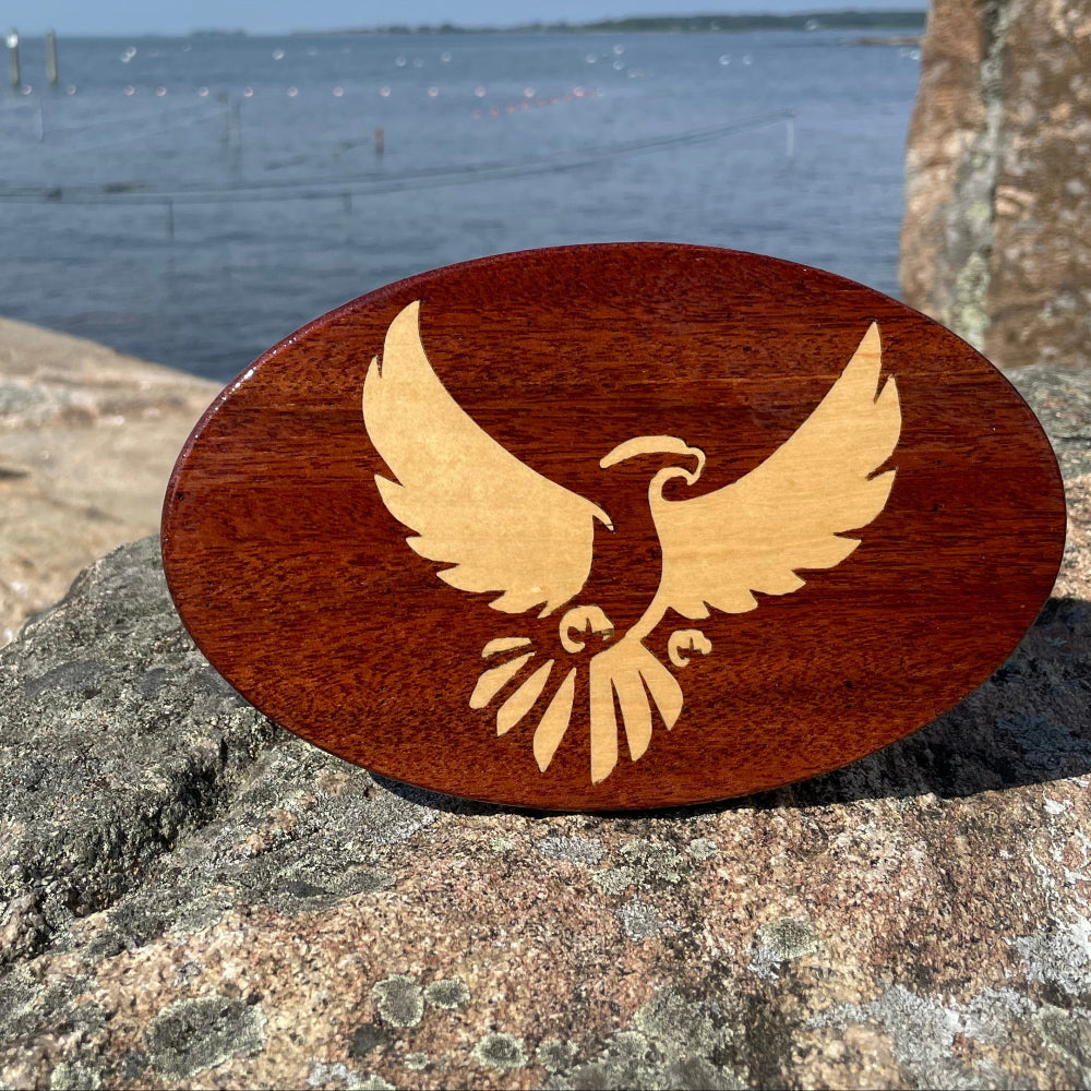 A solid mahogany wooden hitch cover with a maple inlay in the shape of an eagle, placed on a rock near a body of water and a dock.