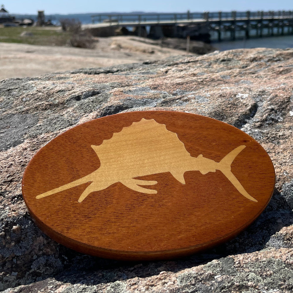 A wooden hitch cover with a sailfish design, placed on a rocky surface with a blurred background.