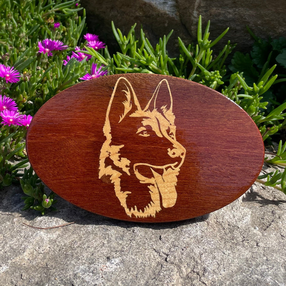 Mahogany hitch cover with a maple inlay featuring a German Shepherd design, displayed on a rock with flowers in the background.
