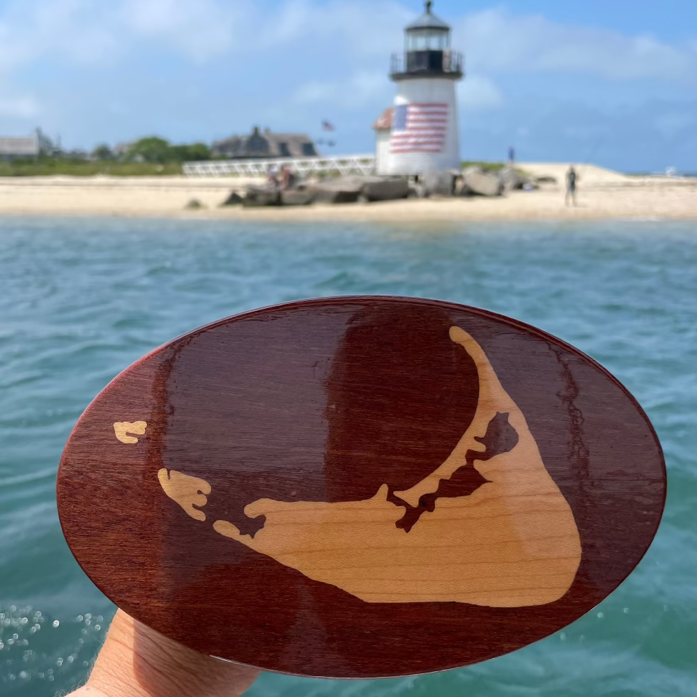 A wooden hitch cover with a Nantucket Island design being held up with a lighthouse in the background.