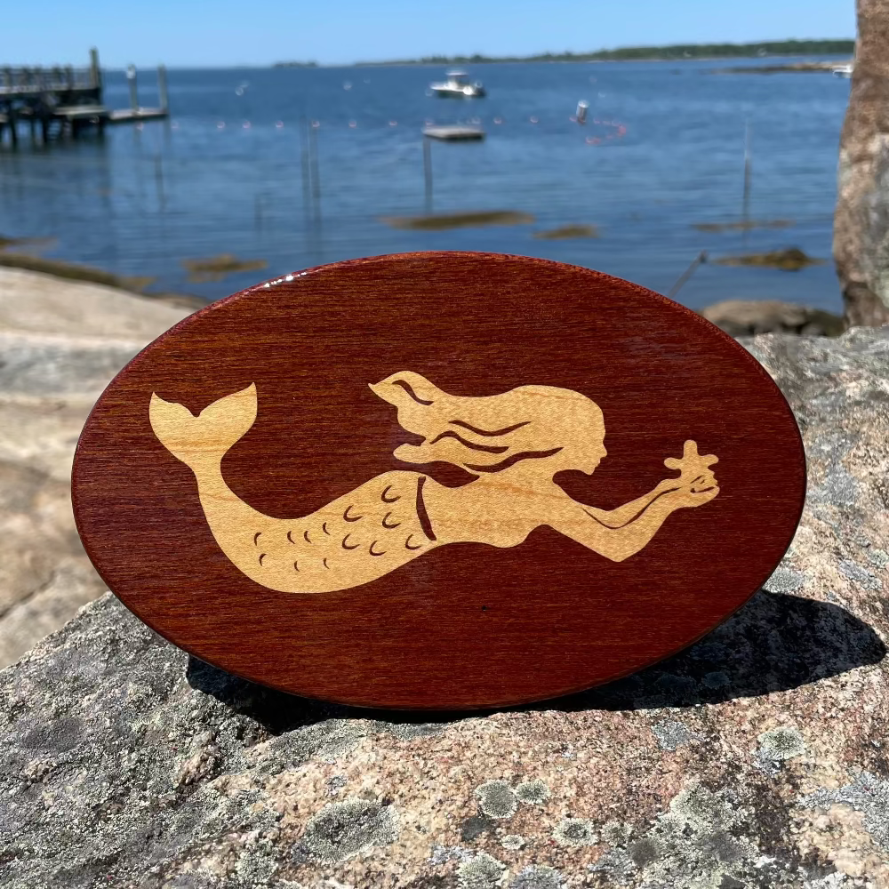 A wooden hitch cover with a mermaid design, placed on a rocky surface with a body of water and sky in the background.