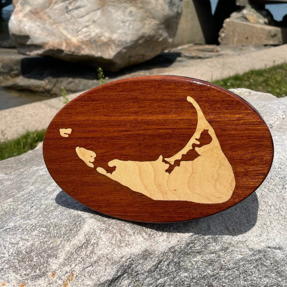 A wooden hitch cover with a Nantucket Island design placed on a rock, with a body of water and a wooden structure in the background.