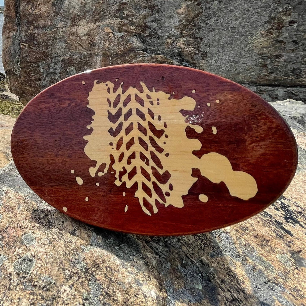 A wooden hitch cover with a Nutmeg Jeepers Puddle Track design, placed on a rocky surface with a body of water and rocks in the background.