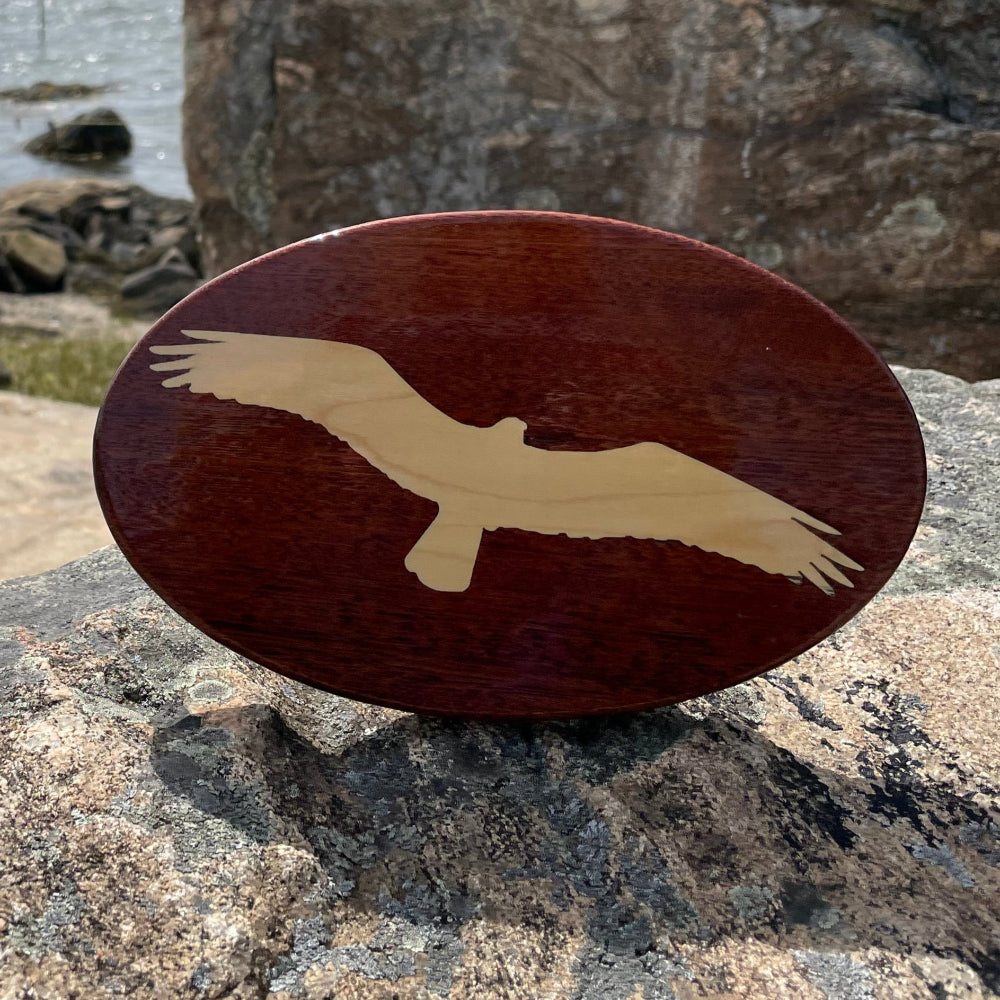 A wooden hitch cover with an osprey design, placed on a rocky surface with a body of water and sky in the background.