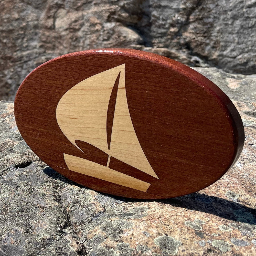 A wooden hitch-cover with a sailboat design, placed on a rock near a body of water and a clear blue sky.
