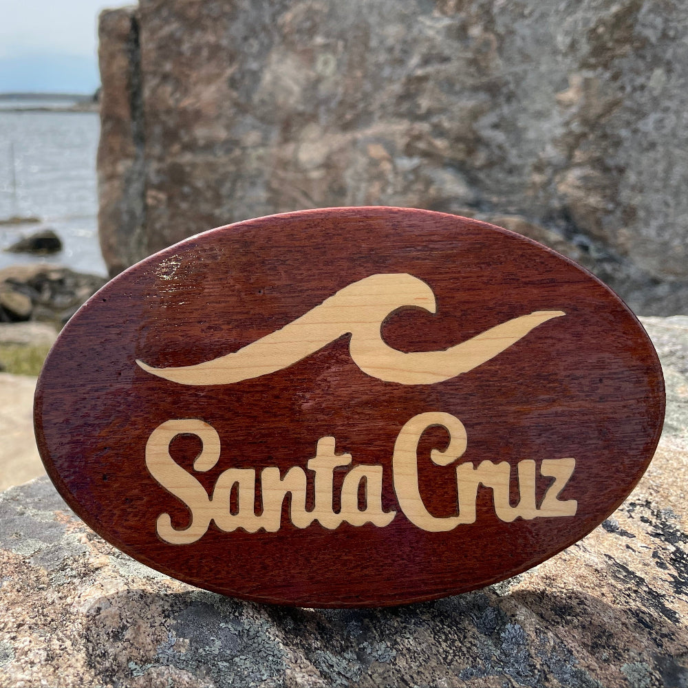 A wooden hitch cover with 'Santa Cruz' text and a wave design, placed on a rocky surface with a body of water and sky in the background.
