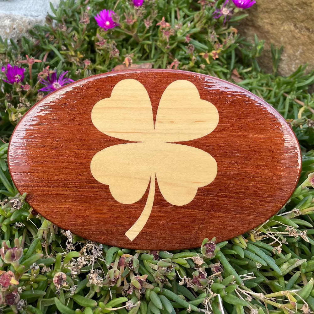 An artisan-crafted wooden hitch cover with a Shamrock design, placed on a bed of flowers.
