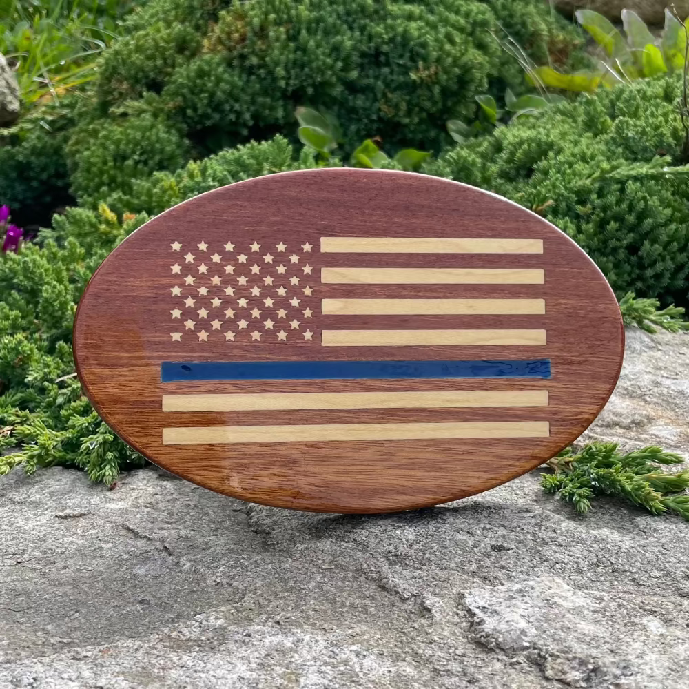 wooden hitch cover with inlay US flag with one stripe as thin blue line balance on a rock with green bush behind it