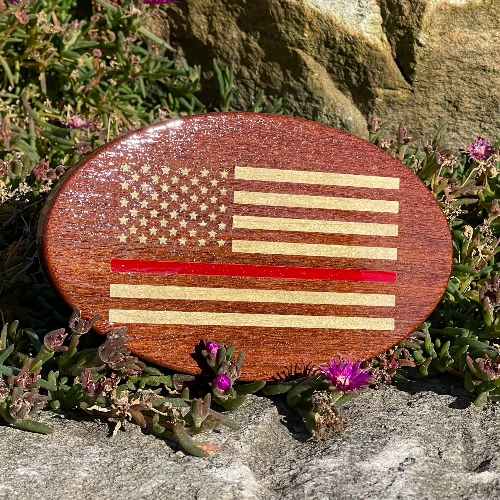 A solid mahogany hitch cover with a maple inlay design featuring an American flag with a thin red line, placed on a rocky ground with flowers behind it.