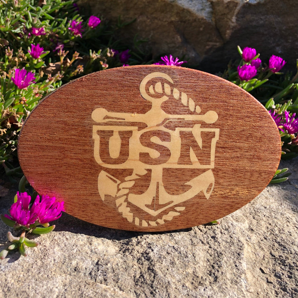 A wooden hitch cover with 'USN' and an anchor emblem engraved, placed on a rock with purple flowers around it.