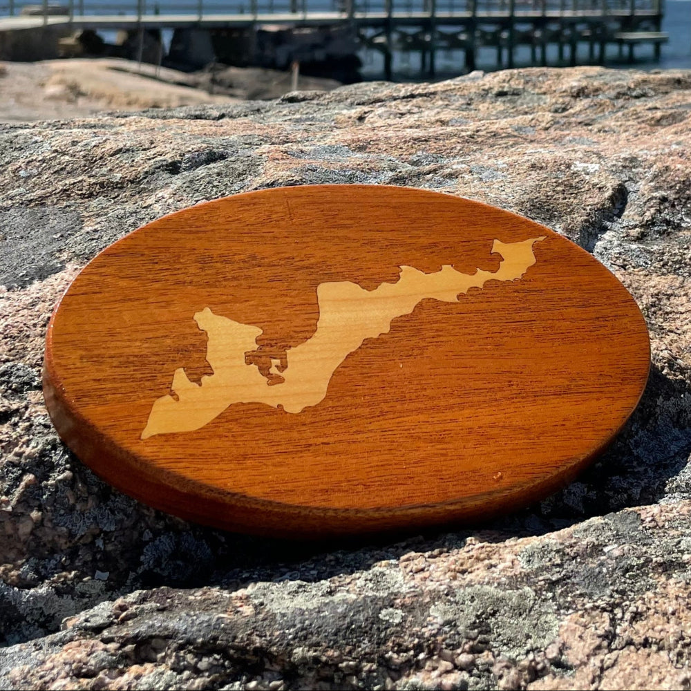 A wooden hitch cover with a map of Fisher's Island design, placed on a rocky surface with a clear sky and water in the background.