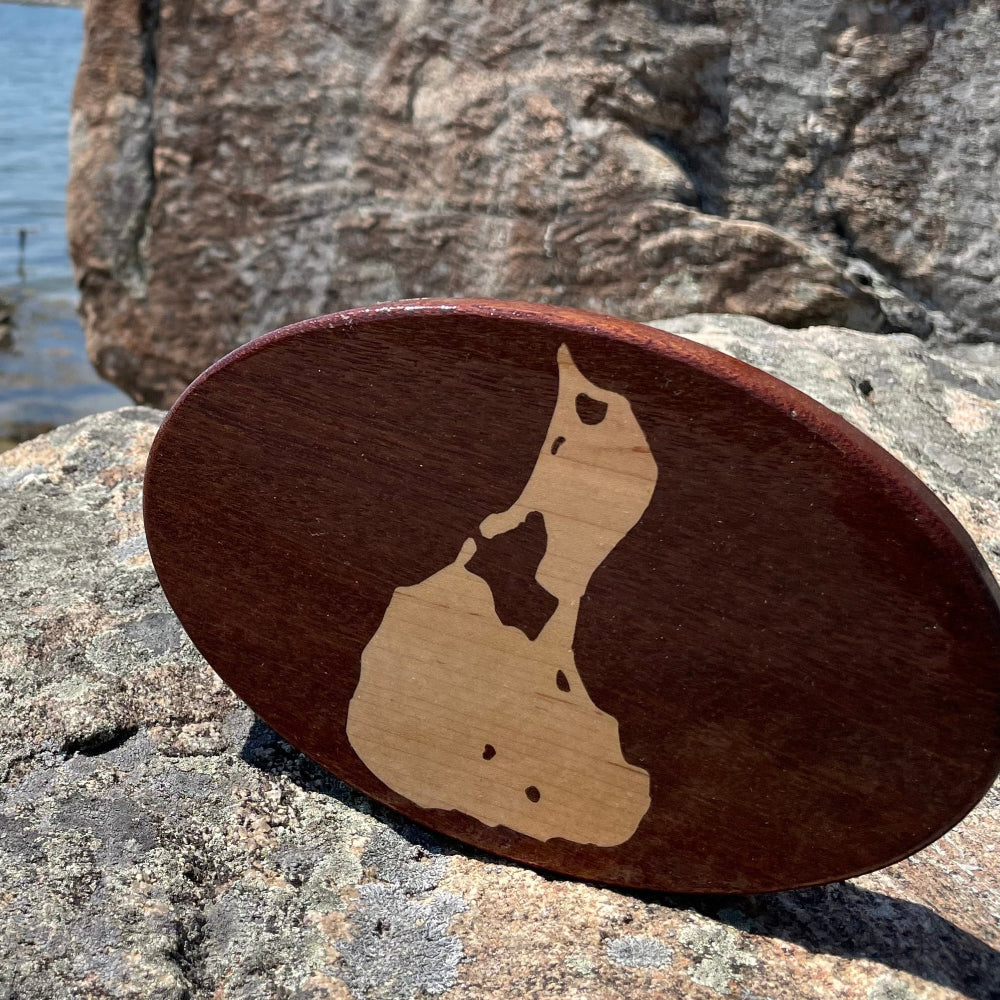 A wooden hitch cover with a maple inlay, designed to celebrate Block Island, resting on a rocky surface with water and sky in the background.