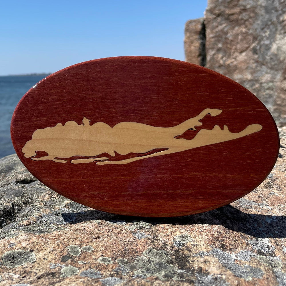 A wooden hitch cover with a Long Island design, placed on a rocky surface with a body of water and sky in the background.