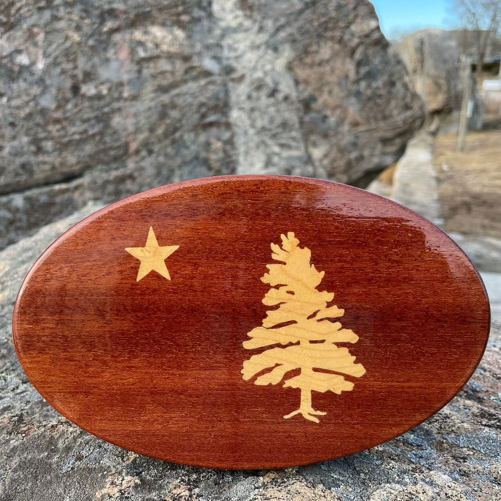 An oval-shaped wooden hitch cover with a Maine flag design, featuring a maple inlay tree and star, placed on a rocky surface.