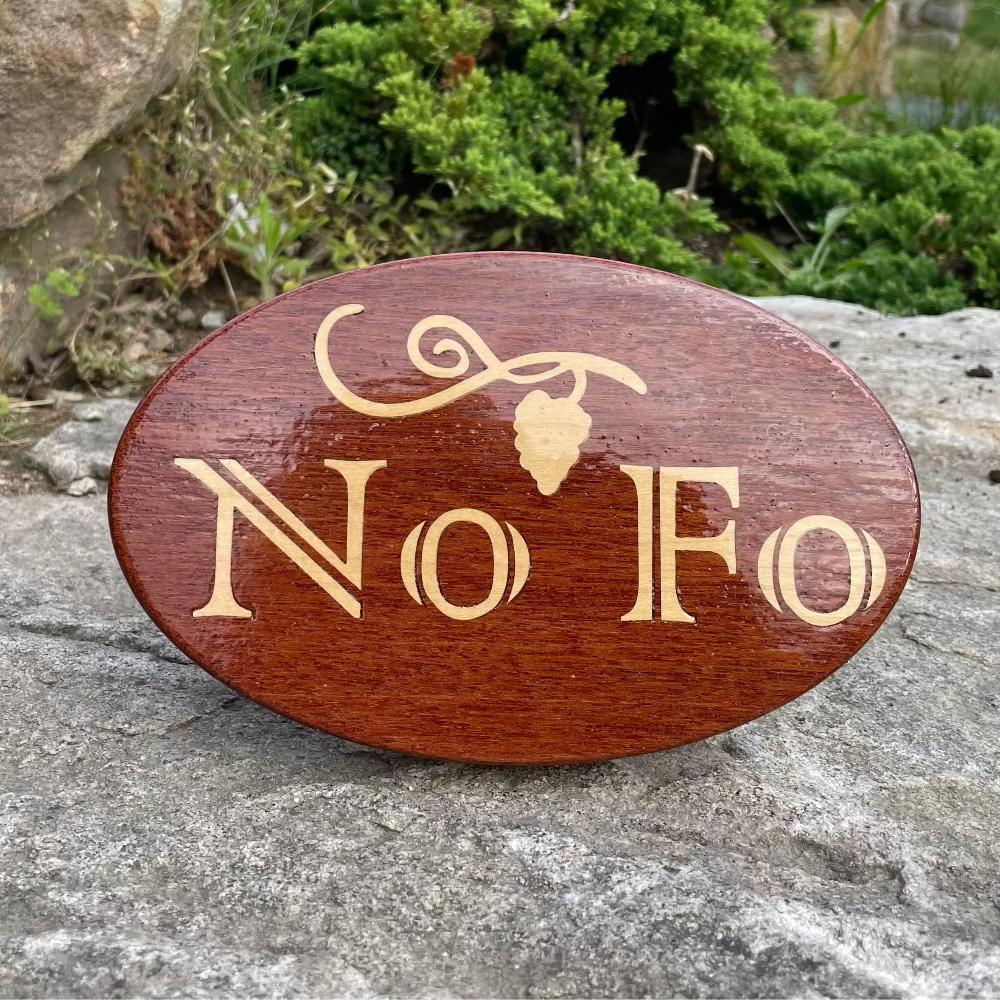A brown mahogany hitch cover with 'NoFo' inlaid and a leaf design, placed on a rocky surface.