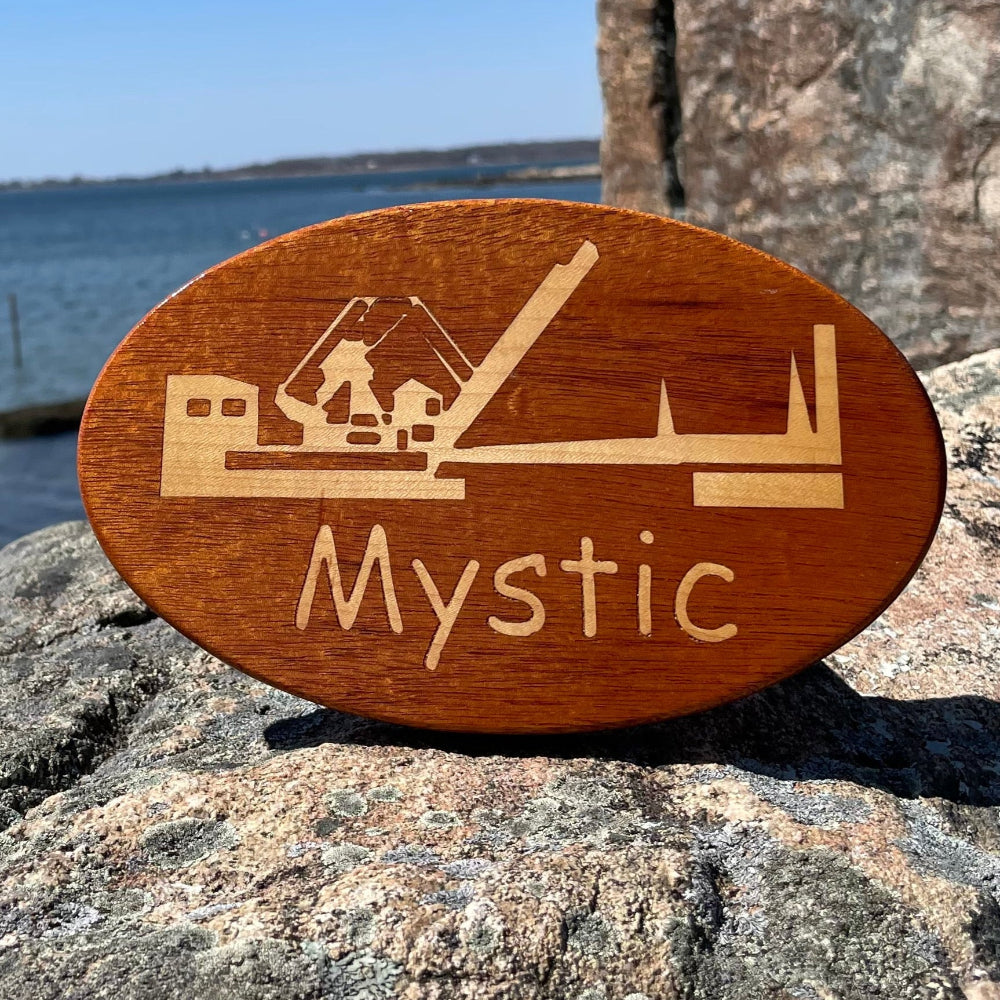 A wooden hitch cover with a Mystic Drawbridge design, placed on a rock near a body of water.