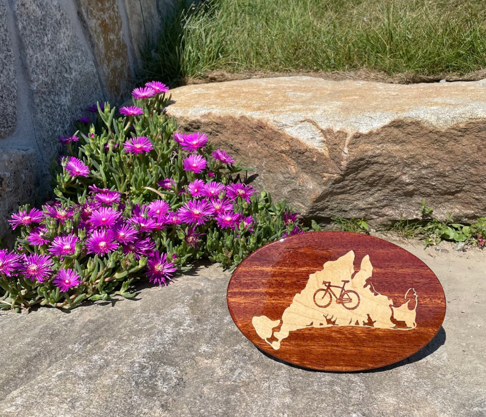 A wooden hitch cover with a maple inlay, designed with a bicycle and the outline of Martha's Vineyard resting on a rock with purple flowers behind it