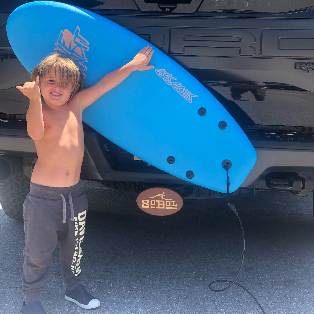 boy oldng a blue surfboard gainst a truck with an inpay wooden hitch cover design in the hitch are a' SOBOL"